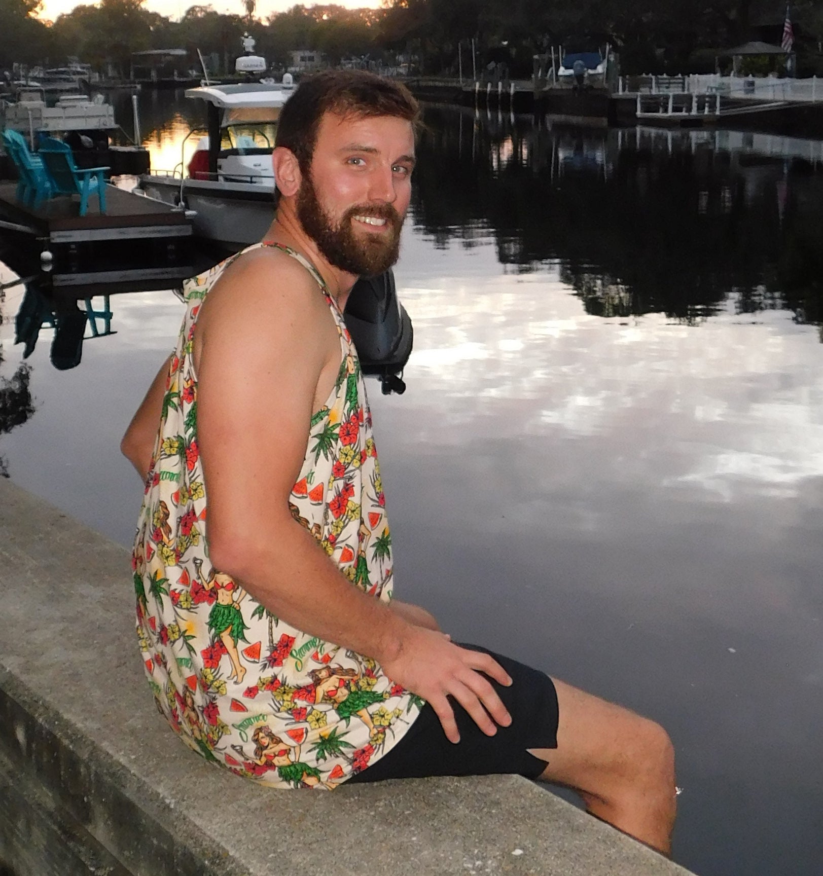a man sitting on a ledge next to a body of water wearing a hula girl hawaiian design tank top 